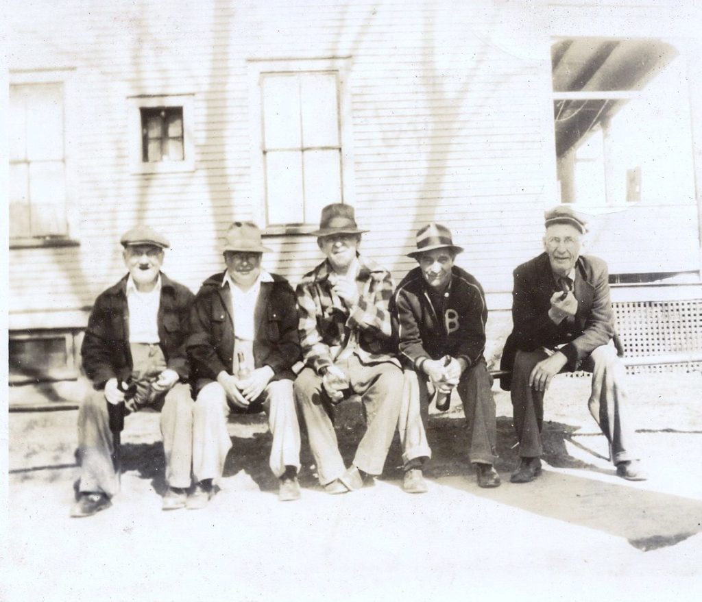 In the foreground there are five older white men sitting on a bench. In the background in front of a building. The photo is black and white.