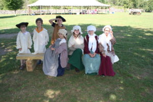 Seven children sit and stand in the shade on a lawn dressed in colonial clothing