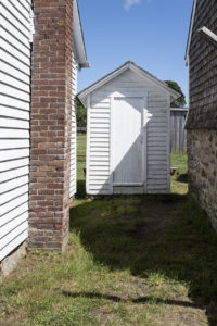 Small white clapboarded shed with a closed door in the narrow alley between two larger outbuildings
