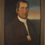Half-length oil portrait of a middle-aged white man with his hand inside a brown jacket. He has a white shirt and dark gray hair. A sunrise sky in the background lights the blue sea, a lighthouse, and a clipper ship.
