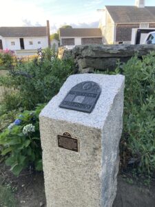 A rectangular granite pillar with a bronze medallion set in the top stands in a garden with a stone wall behind it and barns in the background