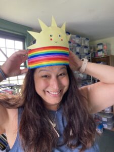 smiling young woman with long, dark hair and olive skin puts a colorful paper rainbow craft hat on her head