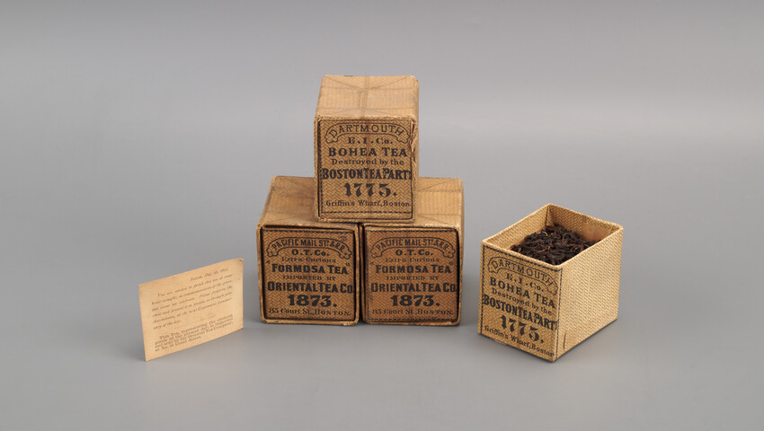 Four commemorative tea boxes. Three are closed and stacked in a pyramid. The fourth is open and its label is visible, showing its origin in 1873 with the Centennial of the Boston Tea Party.