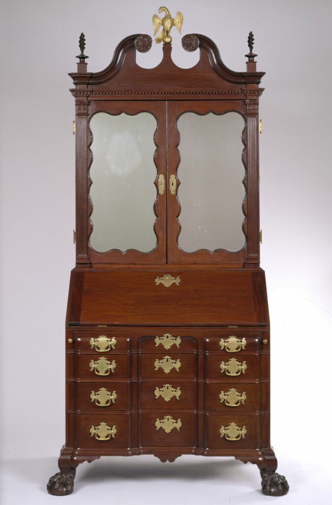 A tall desk with a mirrored cabinet and brass hardware. The top is ornamented with several finials including a brass-colored eagle at the top center.