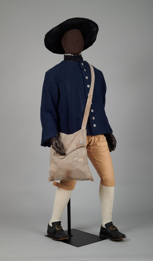A Black mannequin is dressed in a wide-brimmed hat, carefully patched satchel, blue rattan jacket with silver buttons, tan breeches, tall white socks, and buckled shoes. The mannequin is posed in action as if walking forward.