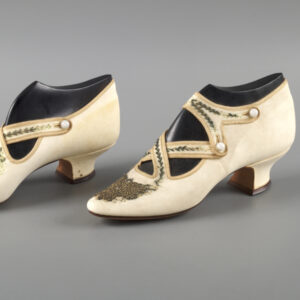 A pair of white leather heels with straps criss-crossing the top of the forms inserted into the shoes. The heels have small buttons fastening the straps, which are decorated with delicate beadwork.
