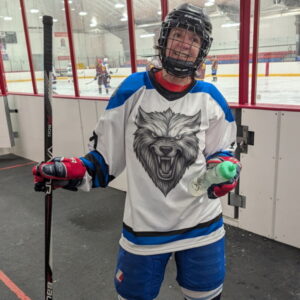 A middle-aged, shorter, White woman with short, curly hair stands wearing a full hockey uniform, including a black helmet with a cage front, in white and blue with a large snarling wolf face on the center of her jersey. She holds a hockey stick in one hand and water bottle in the other. Behind her is an ice rink with hockey players on the ice.
