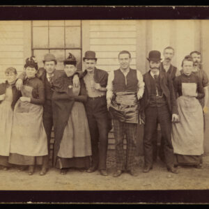 A group of ten men and women in period clothing, including aprons, hats, and waistcoats. They are gathered together for a picture and are close to one another, with arms draped over.
