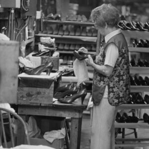 A black and white photo of a woman working in a shoe factory. She stands in the foreground polishing the outside of a pump, with rows of other shoes in the background.