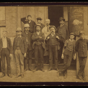 A group of twelve workers stands in front of and on the steps of their factory building, with a sign for "Hawkings, manufacturer of ladies' fine boots" visible. Another worker is visible at the lower right peering out of a basement window.