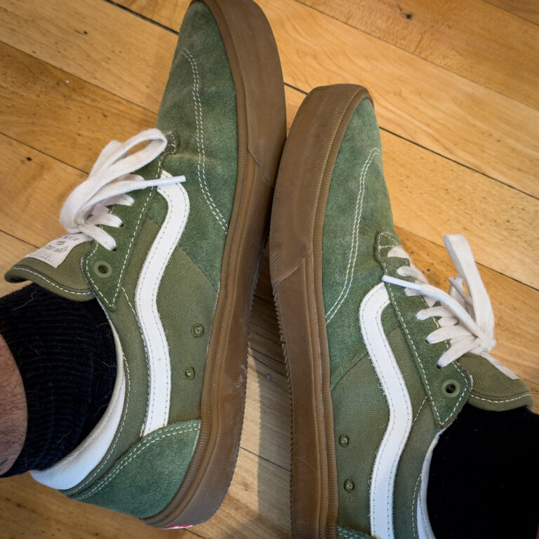 Vans, 2026. An overhead view of a pair of green, white, and brown sneakers worn by a person with black socks. The laces are white and the soles are brown.